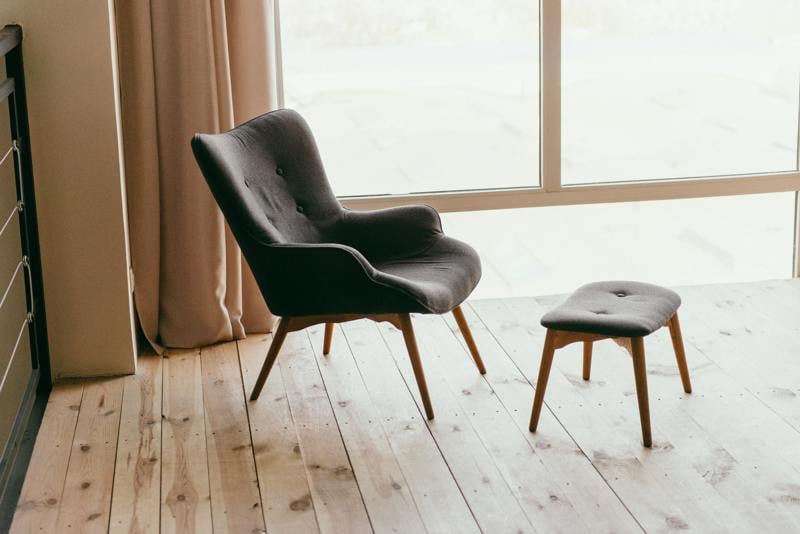 En elegant mörkgrå fåtölj med träben och matchande fotpall står på ett ljust trägolv, placerad vid ett stort fönster med utsikt och ljusa gardiner som ramar in rummet.