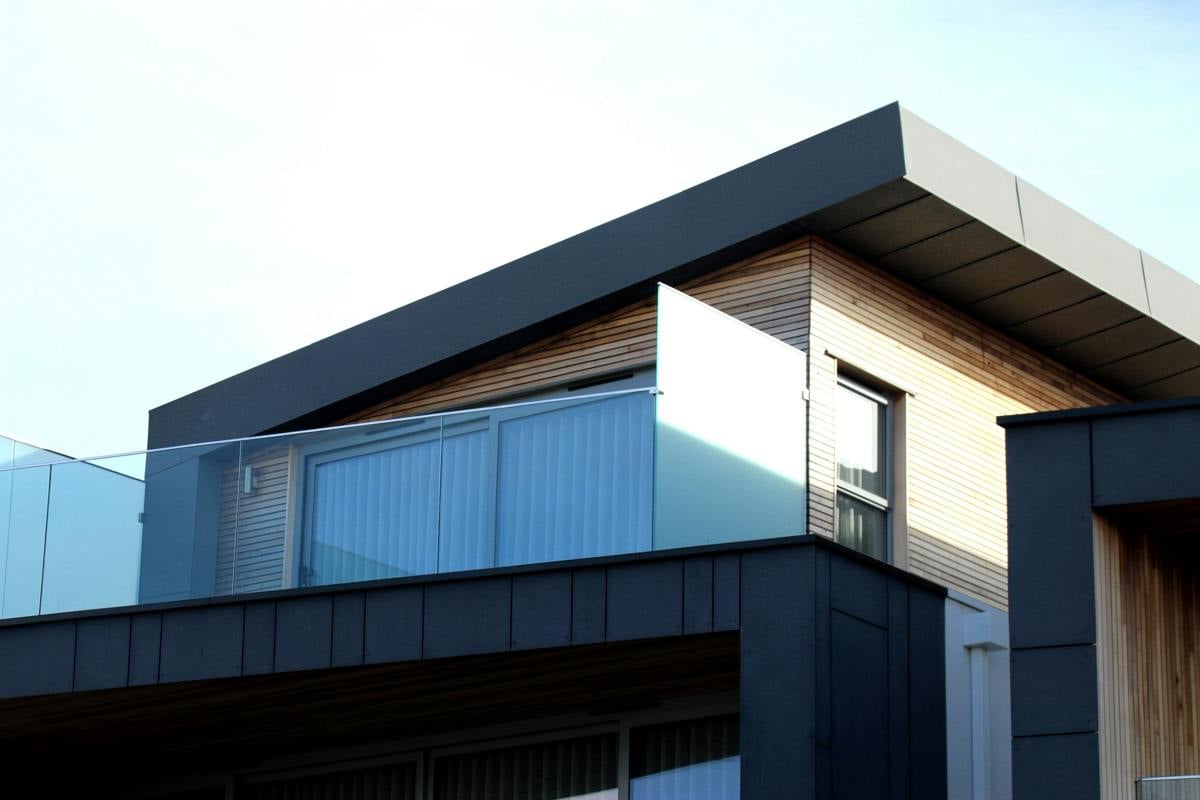 Närbild på en modern bostadsbyggnad med stilren arkitektur, kombinerad träpanel och mörk fasadbeklädnad. En balkong med glasräcke och skjutdörrar ger en ljus och öppen känsla.
