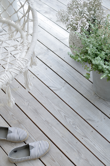 Grått trädäck med en korgstol och randiga tygskor. Två krukor med vita blommor och gröna växter.