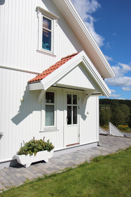 Vitt hus med rött tegeltak, vit dörr med glasrutor, blomsterrabatt framför, grå stengång och gräsmatta.