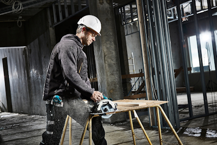 Person med hjälm och skyddskläder sågar plankor med en cirkelsåg i ett byggnadsområde inomhus.