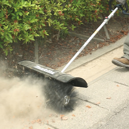 Lövblåsare som sopar bort damm och löv från trottoaren, omgiven av grönska.