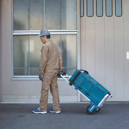 Person klädd i arbetskläder drar en blå verktygsvagn med handtag intill en byggnad.