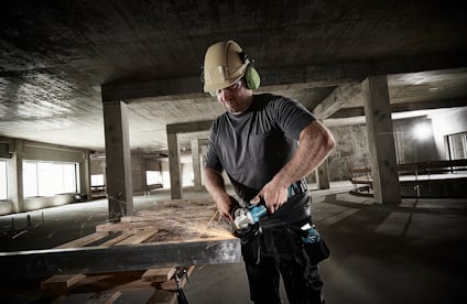 Person i skyddshjälm och hörselskydd slipar metallbalk i en betongbyggnad.