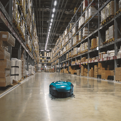 Robotdammsugare med texten "Makita" på en gång i ett lager fullt av hyllor med lådor och förpackningar.