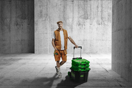 Person i arbetskläder står stilla och håller i en transportkärra med en svart back och tre gröna väskor i ett grått betongrum.