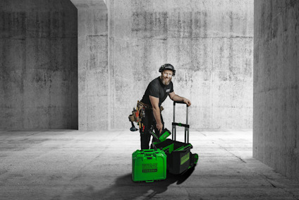 Person med hörselskydd och verktygsbälte lutar sig över en transportkärra och håller en spikpistol. Två gröna väskor står framför en svart back på kärran.