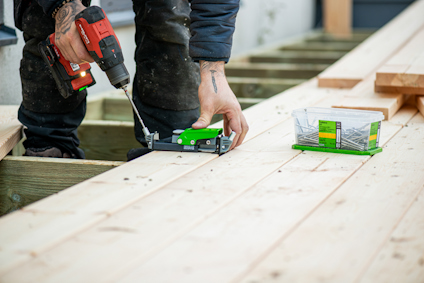 En person lägger en skruv i ett trallmonteringsverktyg för dolt montage och skruvar fast den i en trallbräda med en skruvdragare. En låda med skruvar står bredvid.