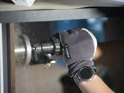 En person borrar i trä med en borrmaskin. De bär grå handskar och en svart klocka.