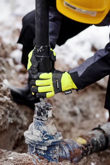 Person med gula handskar och hjälm, arbetar med att vrida ett rör på en byggarbetsplats.