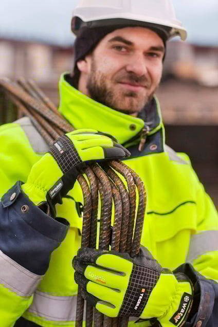 En person i skyddshjälm och reflexjacka håller buntar av armeringsjärn med handskar.
