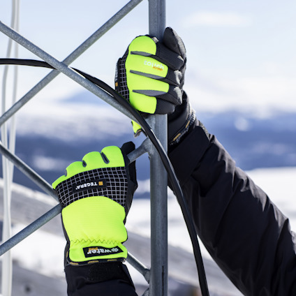 Person med svarta och gula arbetsvantar håller i metallstång utomhus. Snöiga berg i bakgrunden.