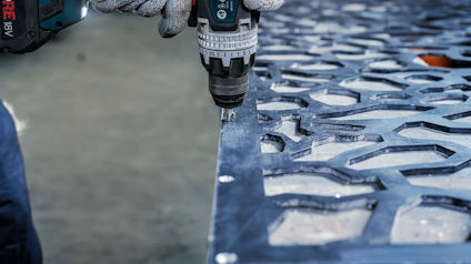 Händer i arbetshandskar håller en borrmaskin som borrar i en metallskiva med geometriskt utskuret mönster.