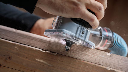 Hand som använder en handöverfräs på en träbit med ett blått dammutsug anslutet.