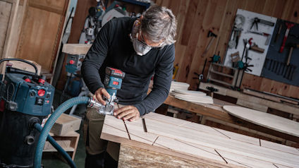 Person med ansiktsmask bearbetar trästycke i verkstad med hjälp av en handhållen maskin.