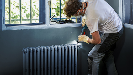 Person i arbetshandskar och skyddsöronsnäckor arbetar med verktyg vid en radiator nära ett fönster.