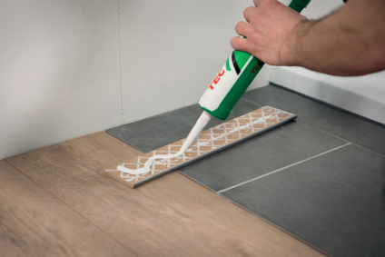 En hand applicerar tätmassa från en tub med munstycke på en bit fogtejp på en golvyta med trä- och kakelplattor.